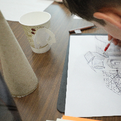 A young student drawing a house on a piece of paper with a cardboard cone and a cup on the table next to the drawing.