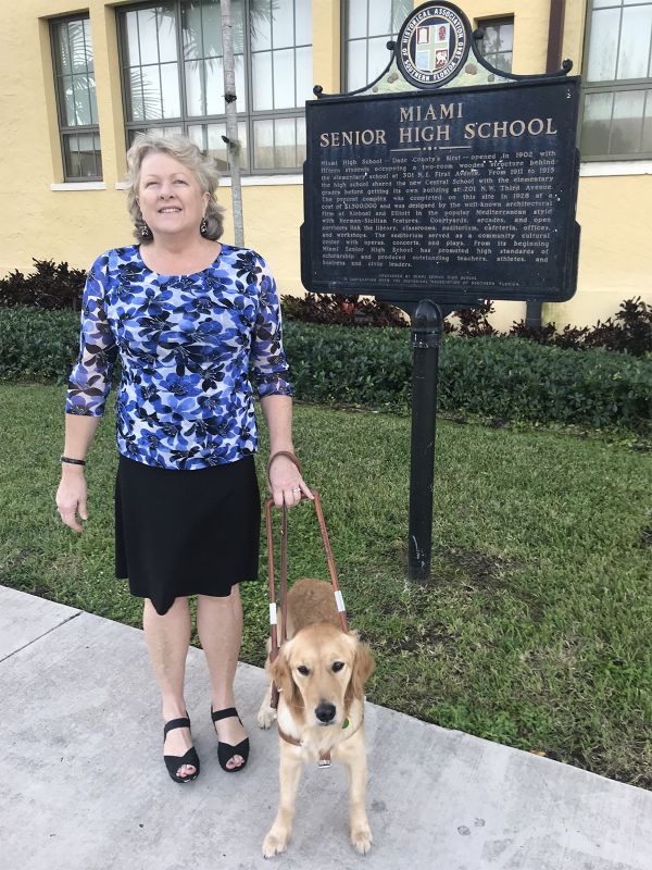 Dr. Janice Bartleson and her guide dog Dusty stand outside next to a Miami Senior High School sign.
