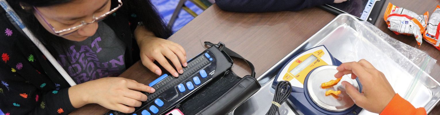 Two blind students weighing cheetos, using a Braille display, and using a tablet during one of our programs for blind teens.