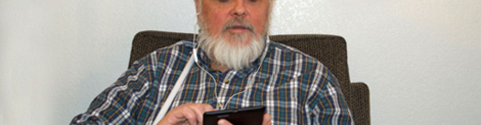A man with a white cane sits and uses a tablet.