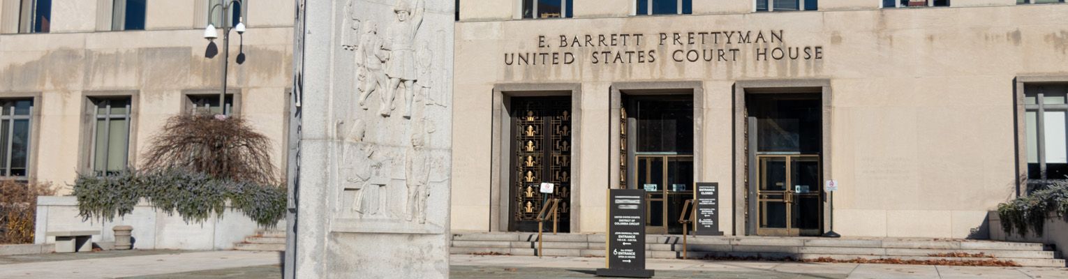 Front of the E Barrett Prettyman United States Federal Courthouse in Washington D.C. The building is tan stone and has three steps leading up to two sets of double doors. There is a stone column in front of the building with engraved images on it. There are signs before and at the top of the steps, and the buildings name is printed in large lettering above the entrance.