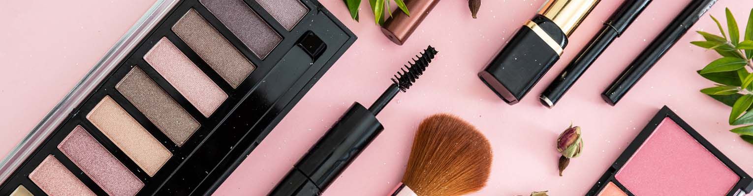 Cosmetics lay spread out on a flat surface. Lipstick, eye shadows and blush, brushes, pencils, and rose buds against pink color background.