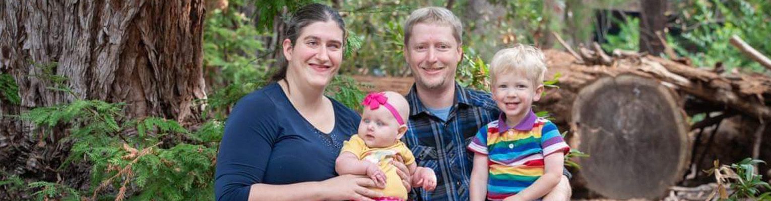 A recent family portrait of Jamie and her family sitting on a log in their favorite redwood grove. Jamie holds her daughter, Selene; her husband, Tim, holds their son, Heleon.