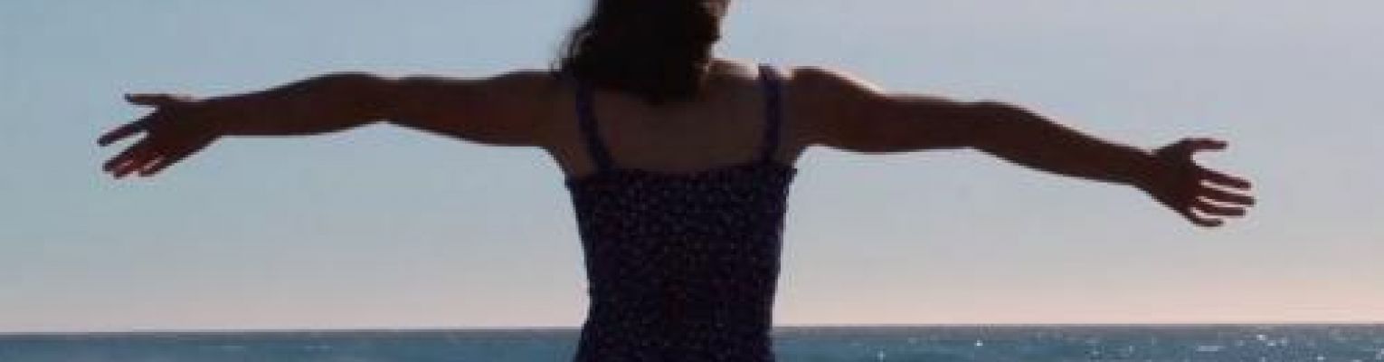 Sarah stands on the beach with her arms outstretched.