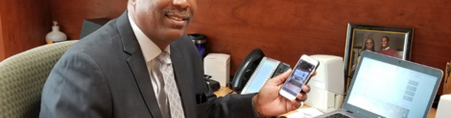 Anil Lewis smiling at his desk holding his iPhone.